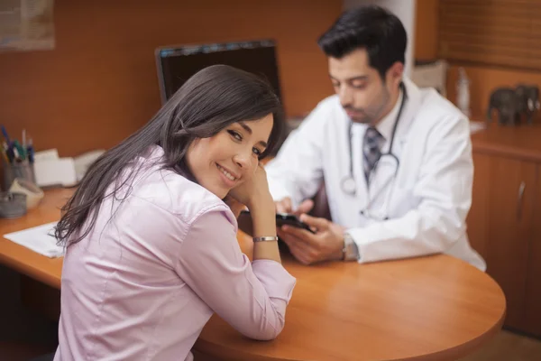 Mujer sonriente sentada en el consultorio del médico en la cita, mirando a la cámara, médico en segundo plano —  Fotos de Stock