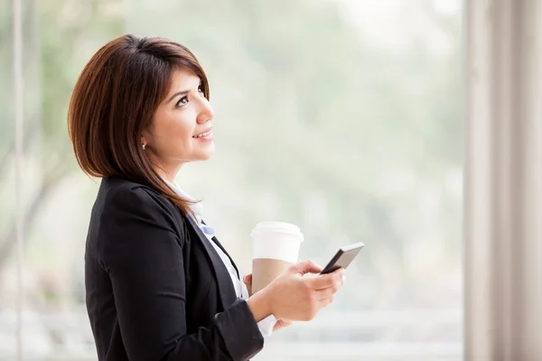 Vista laterale di una bella donna d'affari che scrive sul suo cellulare e tiene in mano una tazza di caffè — Foto Stock