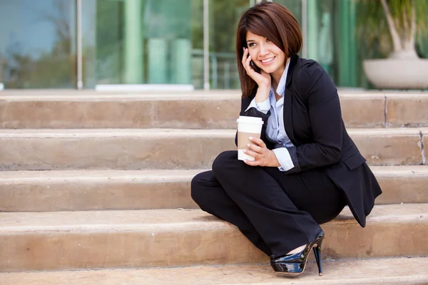 Young smiling business woman sitting on stairway calling with cellphone and holding a cup of coffee — Stock Photo, Image