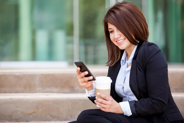 Lächelnde Geschäftsfrau, die auf den Stufen sitzt, Kaffee trinkt und aufs Telefon schaut — Stockfoto