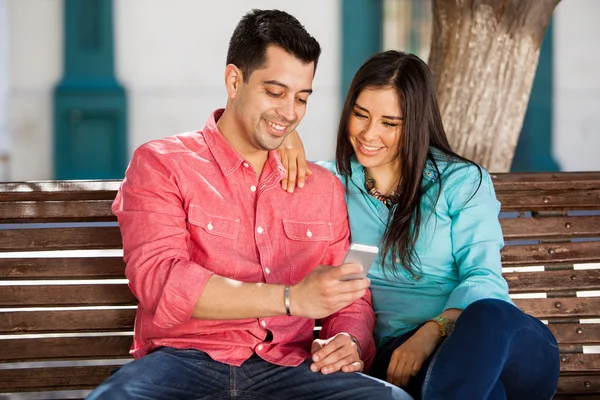 幸せなカップルのベンチに座っていると、スマート フォンを使用して — ストック写真