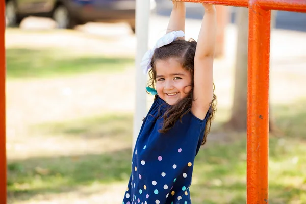 Happy little Hispanic girl hanging from some handlebars in a park — Stock Photo, Image