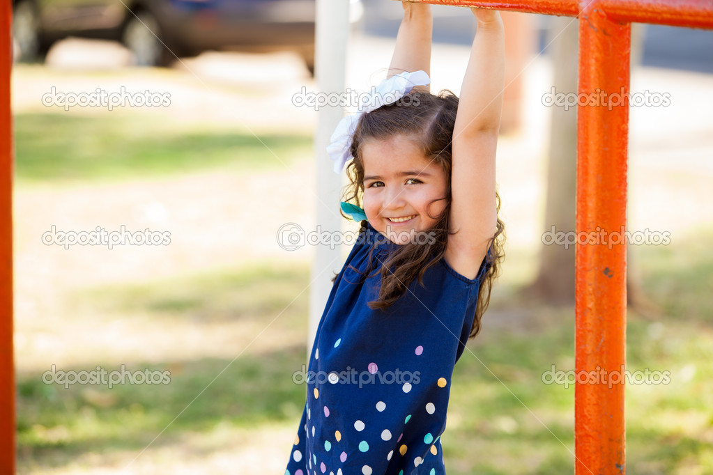 Happy little Hispanic girl hanging from some handlebars in a park