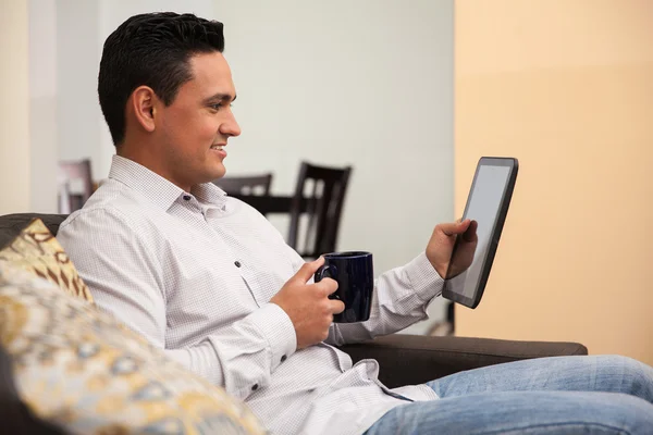 Ontspannen jonge man met een kopje koffie en het lezen van het nieuws op een tablet pc thuis — Stockfoto