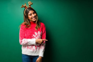 Yeşil bir arka planın önündeki boşluğu gösteren Noel şapkalı neşeli genç kadın.