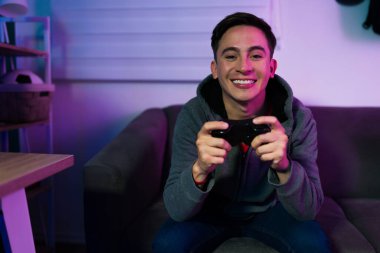 Neşeli genç bir adam, bir video oyunu oynarken tek başına gülümsüyor ve göz teması kuruyor.