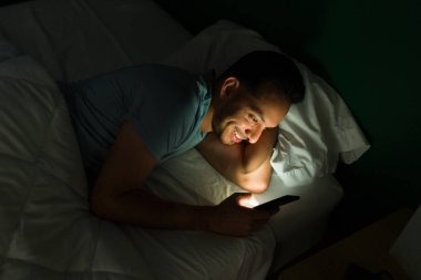 Heyecanlı genç adam gece geç saatlere kadar uyanık kalırken akıllı telefonuyla mesajlaşıyor ve çevrimiçi video izliyor.