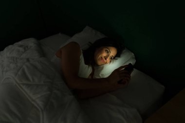 Gece geç saatlere kadar uyanık kalırken akıllı telefonuyla mesajlaşıp video izleyen heyecanlı genç bir kadın.