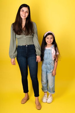 Çekici genç bir anne ve sarı bir stüdyo geçmişiyle göz teması kurarken el ele tutuşan sevimli küçük bir kız.