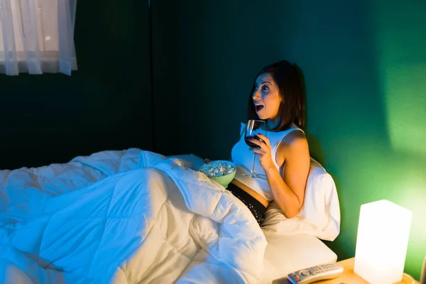 令人惊讶的女人晚上躺在床上一边喝酒一边吃爆米花 一边欣赏一部惊险片 — 图库照片