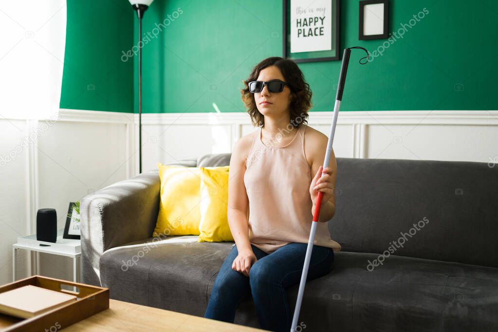 Young woman with a disability sitting on the sofa with a walking cane. Blind woman with black sunglasses waiting at home