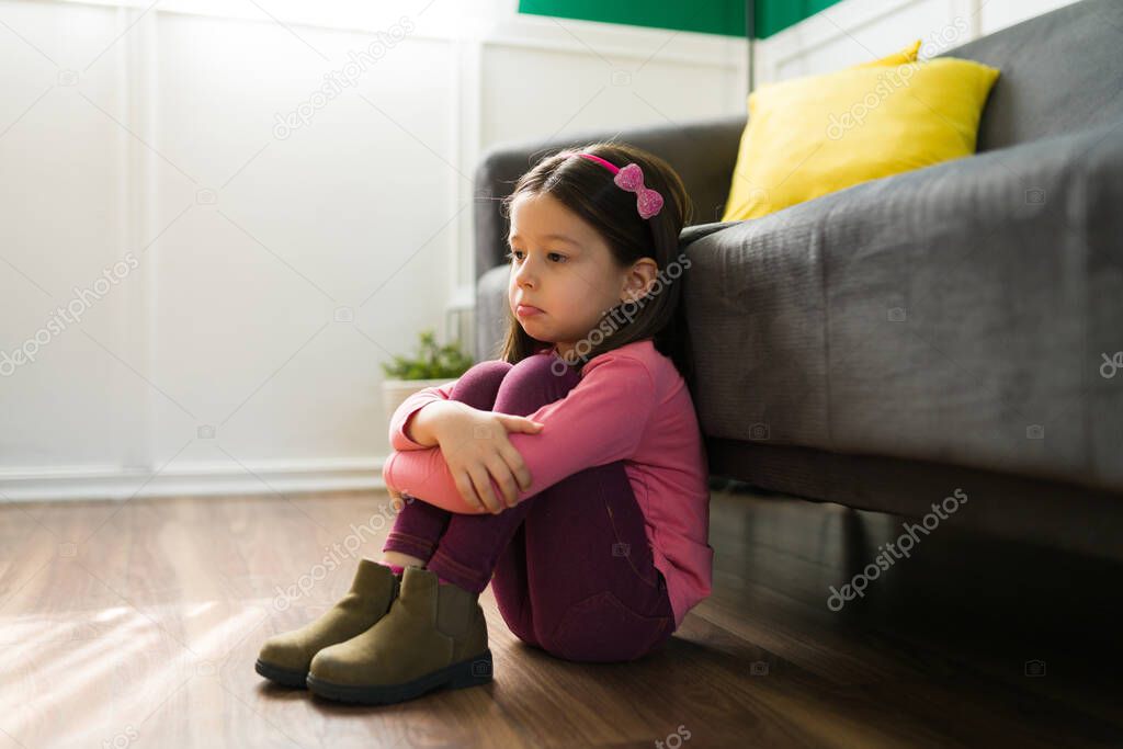 No one wants to play. Depressed young girl hugging her knees and feeling like crying alone at home. Little kid lonely at home