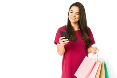Alışveriş merkezinde alışveriş yaparken akıllı telefonuyla mesaj atan neşeli bir kadın. Güzel kadın alışveriş çantalarını taşıyor.