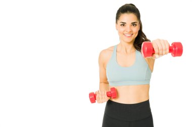 Kol ağırlıklarını kaldırırken gülümseyen, beyaz arka planda spor yapan beyaz bir kadın.