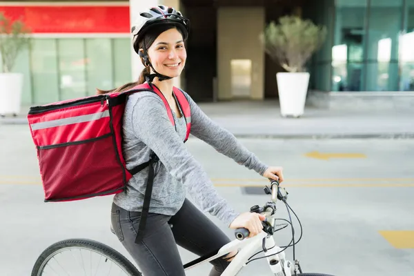 自転車に乗っている間 カメラを見て配達袋を持つラテンの若い女性と食品を提供する作業 — ストック写真