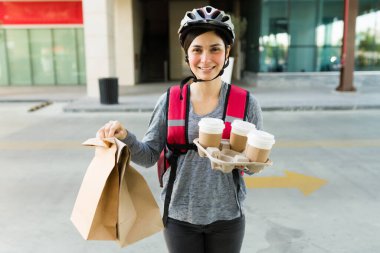 Kahveyle göz teması kurarken gülümseyen mutlu teslimatçı kız ve bir müşteriye yemek paketi götürürken.