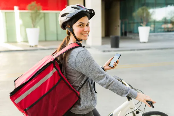 一个快乐的女人 戴着红色的分娩袋 骑着自行车 一边笑着一边用手机看地图 一边送食物 — 图库照片