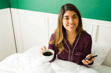 Güzel bir kadın, bir fincan sıcak kahveyle yatakta dinlenirken ve akıllı telefonunda sosyal medya kullanırken sabahtan zevk alıyor.