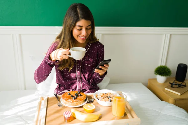 在一个轻松的早晨 美丽的拉丁女人一边喝咖啡 一边在床上吃早餐 一边发短信 — 图库照片
