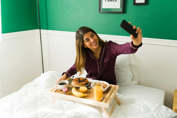 在一个懒洋洋的早晨 一个漂亮的女人在床上吃早餐的时候 拿着智能手机自私自利 — 图库照片
