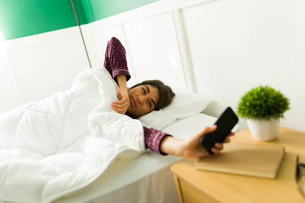 现在几点了 疲惫的年轻女人在床上醒来 看着她在智能手机上的告示 — 图库照片