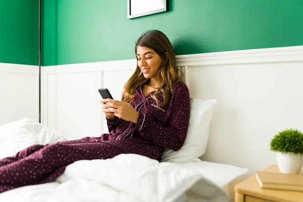早上很慢20多岁的快乐女人坐在床上 一边放松 一边用手机发短信 — 图库照片