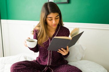 Akıllı genç bayan pijama giyiyor ve yatak odasında kitap okurken bir fincan kahvenin tadını çıkarıyor.