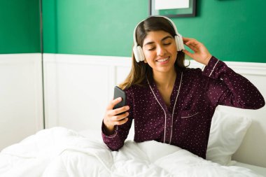 Güzel pijamalı rahat bir Latin kadın yatak odasında dinlenirken kablosuz kulaklıkla müzik dinliyor.