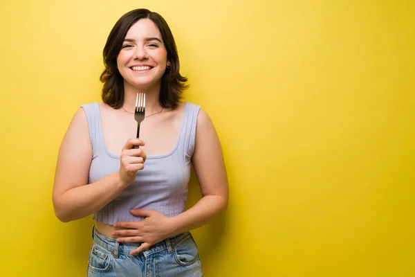 食事の後にフォークを持っている陽気な若い女性と彼女の胃をこすりながら食べ物でいっぱい感じて — ストック写真