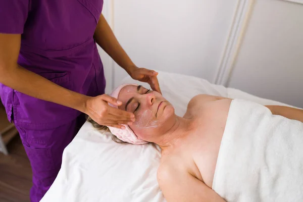 女医生在一位镇静的老年妇女的脸上涂上润肤霜 并在温泉进行面部治疗 — 图库照片
