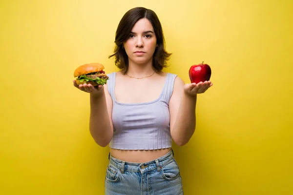 均衡饮食 一个严肃的年轻女人一边拿着一个苹果和一个汉堡包一边看着相机 — 图库照片