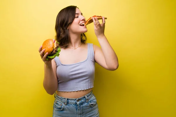 我喜欢披萨 快乐的年轻女人一边吃着一片披萨 一边拿着一个黄色背景的汉堡包 — 图库照片
