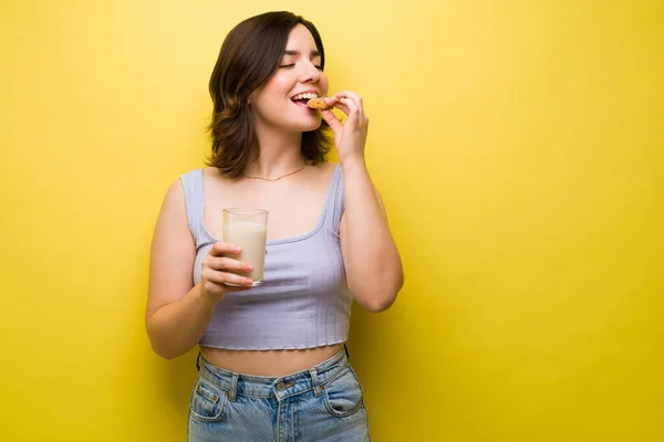 快適な食べ物 魅力的な若い女性食べるクッキーとミルクに対して黄色の背景 — ストック写真