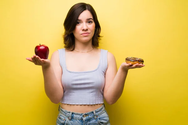我想吃一个甜甜圈 一个女人拿着一个红苹果和甜甜圈 眼神交流 看起来很沮丧 很不高兴 — 图库照片