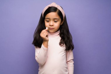 İlacımı almam gerek. Mor bir arka plan önünde elinde hap ya da vitamin tutan hasta bir çocuk.