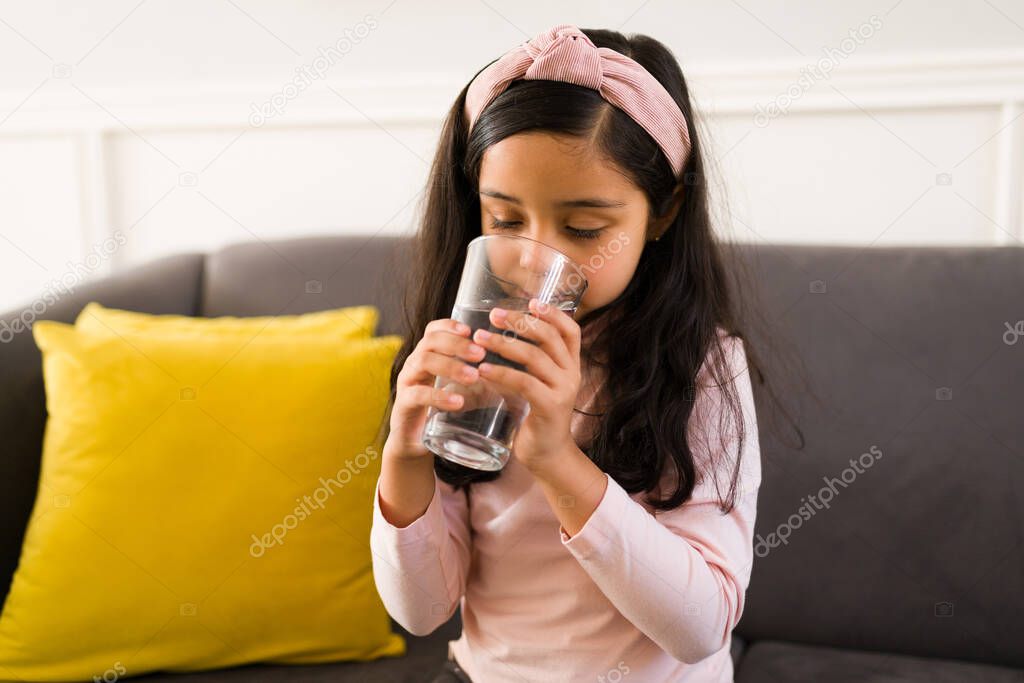 Adorable girl drinking a glass of water while relaxing at home. Hispanic girl feeling thirsty