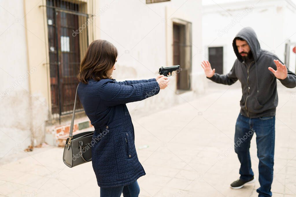 Stop right there! Angry young woman pointing a gun to a criminal thief. Young man and mugger putting his hands up on the street