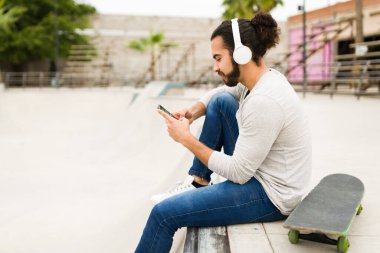 Latin genç adam müzik dinlemekten zevk alıyor. Akıllı telefondan mesaj atıyor ve bir arkadaşının parkta birlikte paten yapmasını bekliyor.