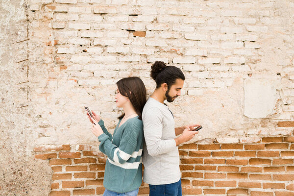 Парень и девушка стоят спиной к спине, переписываясь на смартфонах с кирпичной стеной.