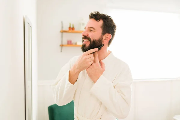 Cheerful young man feeling happy with beard while combing and looking in the mirror before going out