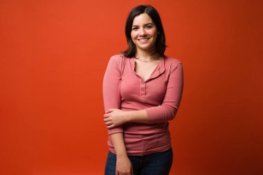 Beyaz bir kadın stüdyo arka planında dikilirken kendine güveniyor ve gülümsüyor.