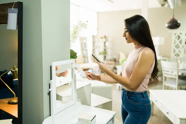 Beautiful young woman looking at the price of a vanity mirror and taking a picture with a smartphone at the furniture store