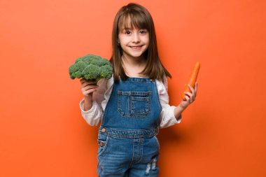 Hoş görünümlü küçük bir kız havuç ve brokoli tutuyor. Bir sürü sağlıklı yemek yediğini gösteriyor.