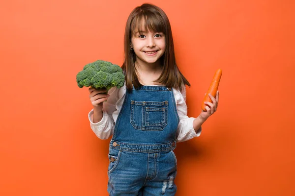 可愛い女の子はカジュアルに身を包んだとニンジンを保持し いくつかのブロッコリーを見るために彼女は健康食品の多くを食べる — ストック写真