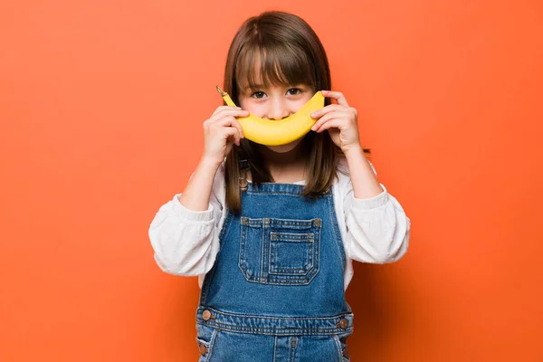 スタジオで果物と楽しみながら彼女の笑顔の代わりにバナナを保持している愛らしい小さな白人の女の子 — ストック写真