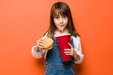 Sıradan giyinmiş küçük bir kızın portresi ve elinde hamburger tutarken büyük bir gazozdan bir yudum içmesi.