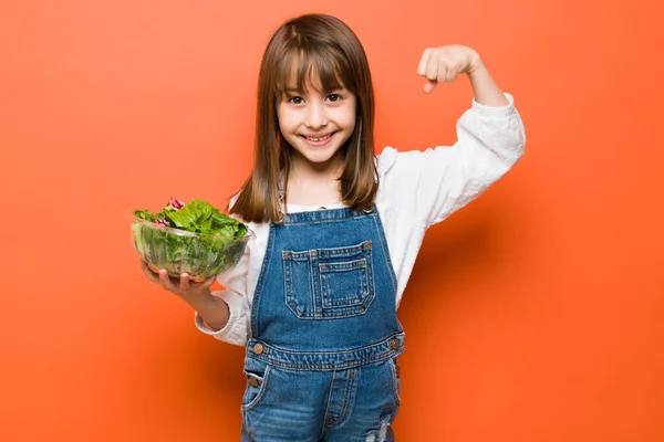 幸せな小さな白人の女の子を保持しているサラダのボウルとショー彼女の強い腕のために健康的な食べ物を食べる — ストック写真
