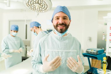 Ameliyata başlamadan önce ameliyat önlüğü ve eldiven giyerek gülümseyen yakışıklı bir cerrah. 