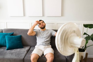 Handsome young man cooling with a fan on. Latin man sweating putting a cold lemonade glass on his forehead 