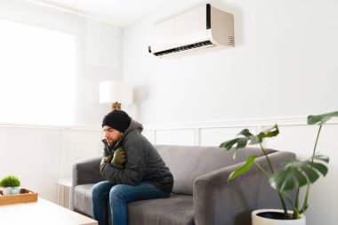 Cold latin man sitting on the sofa enjoying the freezing air while the AC unit is on in the living room